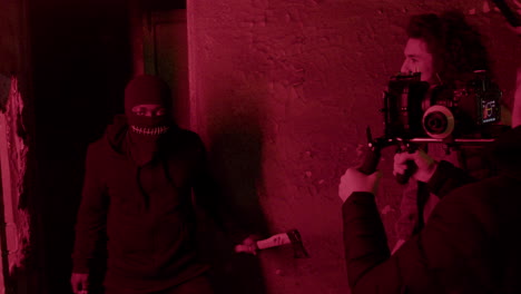 Cameraman-And-Production-Worker-Recording-A-Scene-Of-An-Man-In-Black-Balaclava-Holding-An-Ax-In-A-Ruined-Building-1