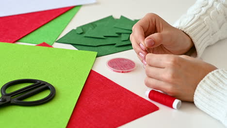 Female-hands-get-sewing-thread-in-needle-to-make-Christmas-crafts-with-colored-fabrics