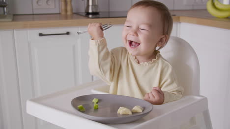Cute-baby-girl-eating-avocado-slice-using-fork-while-sitting-in-high-chair-in-the-kitchen