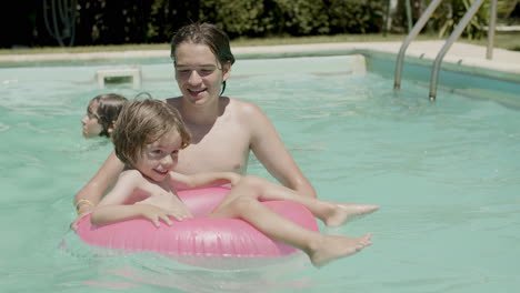 Cheerful-little-boy-floating-on-inflatable-ring-in-swimming-pool-with-the-help-of-his-brother