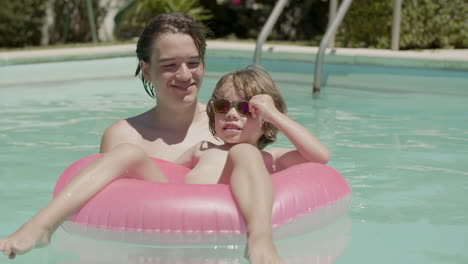 Cheerful-little-boy-floating-on-inflatable-ring-in-swimming-pool-with-the-help-of-his-brother-and-posing-for-camera