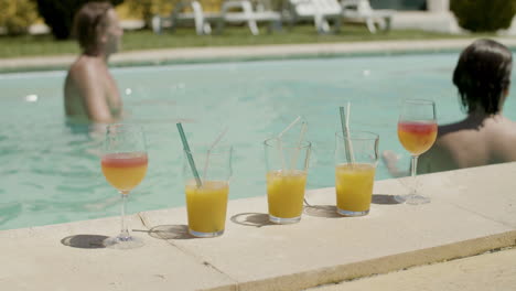 Nahaufnahme-Von-Orangensäften-Und-Cocktails-Am-Pool,-Während-Im-Hintergrund-Eine-Glückliche-Familie-Mit-Ball-Im-Schwimmbad-Spielt
