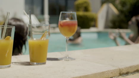 Close-up-of-orange-juices-and-cocktails-on-poolside-while-in-the-background-a-happy-family-playing-with-ball-in-swimming-pool