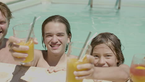 Happy-family-in-swimming-pool-with-their-cocktails-on-poolside-looking-at-the-camera-and-smiling
