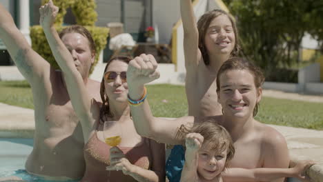 Happy-Caucasian-family-standing-in-swimming-pool-and-raising-their-arms-up-while-looking-at-the-camera