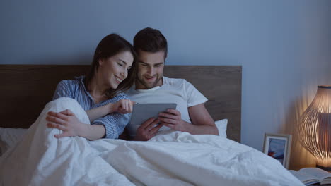 Young-caucasian-couple-sitting-the-bed-and-watching-a-video-on-the-tablet-device-at-night