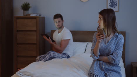 Good-looking-just-married-man-and-woman-in-pajamas-scrolling-and-typing-on-their-smartphones-while-sitting-on-the-different-corners-of-the-bed-at-night