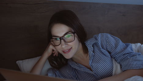 Close-up-view-of-smiling-woman-using-laptop-and-smiling-while-watching-something-in-the-evening-in-her-bed