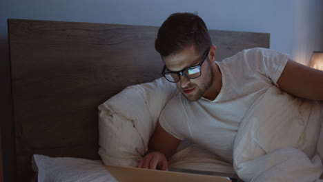 Close-up-view-of-smiling-man-using-laptop-and-smiling-while-watching-something-in-the-evening-in-her-bed