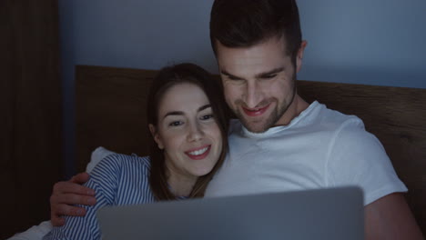 Smiling-good-looking-Caucasian-young-couple-watching-something-funny-on-the-laptop-in-the-bed-at-night