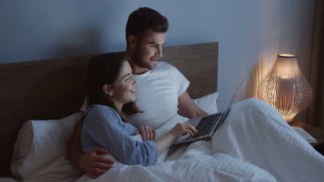 smiling-good-looking-Caucasian-young-couple-watching-something-funny-on-the-laptop-in-the-bed-at-night
