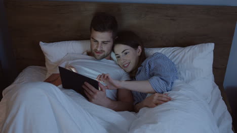 Top-view-of-smiling-Caucasian-wife-and-husband-lying-in-the-bed-using-tablet-under-blanket-late-in-the-evening