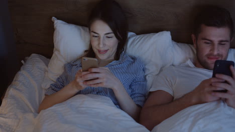 Top-view-of-young-Caucasian-woman-and-man-lying-in-the-bed,-chatting-and-laughing-on-the-smartphone-late-in-the-night