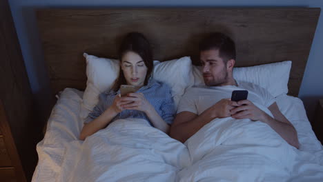 Top-view-on-the-just-married-Caucasian-couple-after-a-quarrel-lying-in-the-bed-at-night-and-typing-on-the-smartphones,-then-they-look-at-each-other