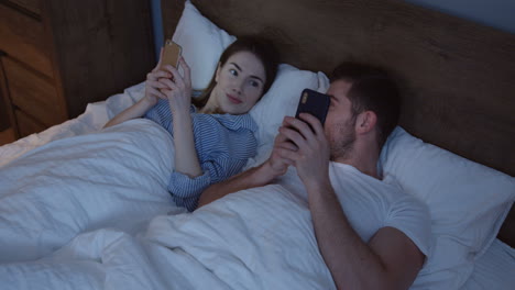 Young-smiling-Caucasian-man-and-woman-lying-in-the-bed-in-the-evening-and-playing-on-smartphones,-then-they-turn-to-each-other