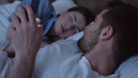 Close-up-view-of-young-Caucasian-man-lying-in-the-bed,-chatting-on-the-smartphone-late-in-the-night-and-looking-at-his-wife-who-sleeping-beside-him