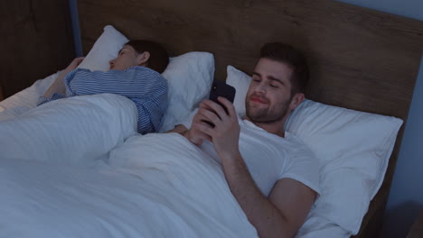 Young-smiling-Caucasian-man-lying-in-the-bed-near-his-young-wife-and-chatting-on-the-smartphone-late-in-the-night