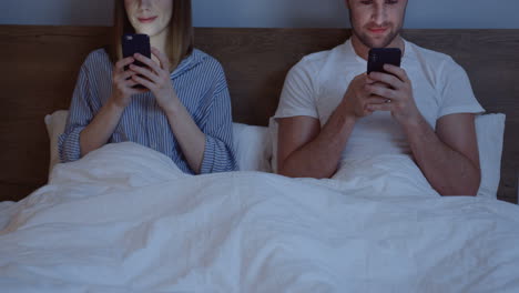 Caucasian-couple-using-smartphones-while-they-are-sitting-in-the-bed-under-the-blanket