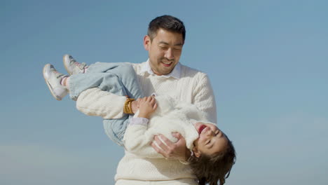 Happy-Japanese-dad-holding-little-daughter-in-his-arms-and-tickling-her-at-the-beach