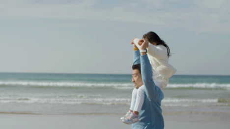 Happy-Japanese-father-walking-along-seashore-with-his-daughter-on-his-shoulders