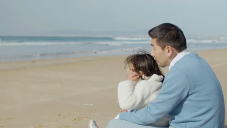 Japanese-dad-and-cute-little-daughter-sitting-at-seashore,-talking-and-enjoying-the-view