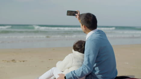 Back-view-of-a-man-holding-smartphone-and-taking-selfie-while-hugging-his-lovely-daughter-at-the-beach