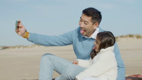 Japanese-dad-and-daughter-taking-selfie-on-mobile-phone-and-showing-tongues-while-spending-time-together-at-the-beach