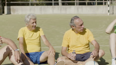 Jugadores-De-Fútbol-Senior-Cansados-Sentados-En-Círculo-En-El-Campo,-Hablando-Juntos-Y-Bebiendo-Agua