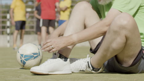 Sportive-senior-football-player-sitting-on-pitch-and-lacing-sneakers