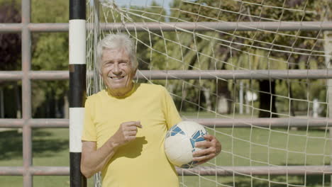 Cheerful-elderly-sportsman-standing-next-to-goalpost,-holding-ball-and-looking-at-the-camera