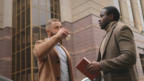 Bottom-view-of-caucasian-and-african-american-businessman-in-elegant-clothes-talking-in-the-street-in-autumn-while-one-of-them-drinking-coffee-and-the-other-holding-a-smartphone