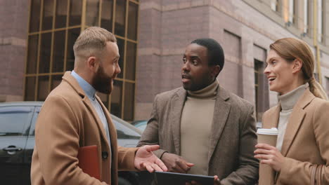 Two-caucasian-and-african-businessman-and-caucasian-businesswoman-talking-about-their-work-in-the-street-in-autumn-while-using-a-tablet