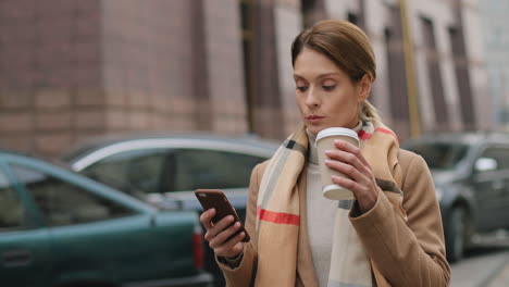 Caucasian-charming-businesswoman-wearing-coat-and-scarf-walking-on-the-street-while-drinking-hot-coffee-and-texting-on-the-smartphone