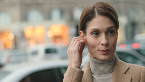 Close-up-view-of-caucasian-businesswoman-wearing-wireless-headphones-while-talking-on-the-phone-and-drinking-coffee-in-the-street-in-autumn