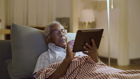 Anciano-Negro-Acostado-En-La-Cama-Navegando-Por-La-Red-Usando-Una-Tableta-Digital