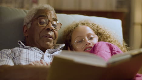 Cute-girl-and-her-grandpa-lying-on-couch-reading-book-together