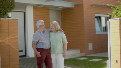 Happy-senior-couple-standing-next-to-new-house,-hugging-and-talking,-then-they-smiling-at-the-camera
