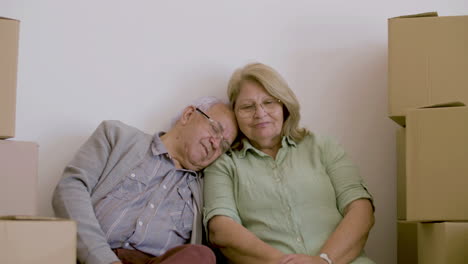 Front-view-of-tired-and-cheerful-elderly-wife-and-husband-sitting-on-floor-with-carton-boxes-after-moving
