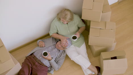 Tired-senior-couple-lying-on-floor-and-drinking-tea-after-moving