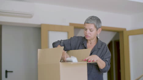 Mature-woman-unpacking-carton-boxes-after-moving-to-new-house