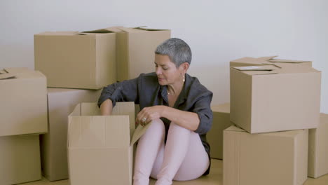 Cheerful-woman-sitting-on-floor-and-unpacking-cardboard-boxes