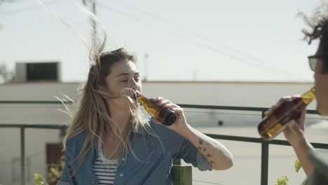 Happy-woman-sitting-on-rooftop-and-clinking-bottles-of-beer-with-a-friend-on-a-windy-day
