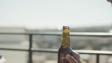 Close-up-shot-of-two-unrecognizable-friends-clinking-bottles-of-beer-while-having-party-on-rooftop