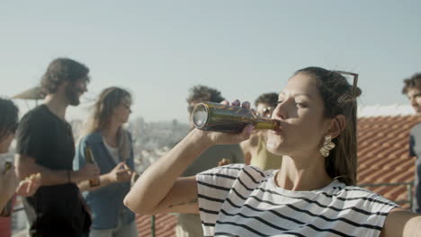 Portrait-of-a-young-Caucasian-woman-looking-at-the-camera-and-drinking-beer-while-having-a-rooftop-party-with-friends