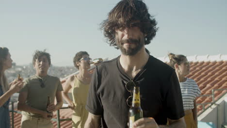 Portrait-of-a-bearded-Caucasian-man-looking-at-the-camera-and-drinking-beer-while-having-a-rooftop-party-with-friends