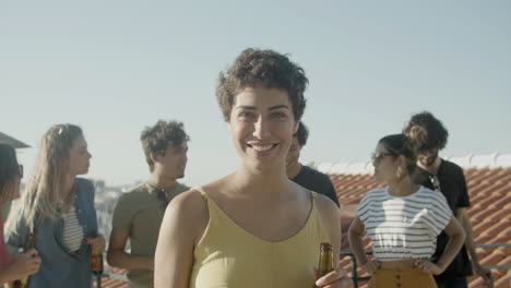 Portrait-of-a-happy-Caucasian-woman-with-short-hair-holding-a-beer-and-looking-at-the-camera-while-having-a-rooftop-party-with-friends
