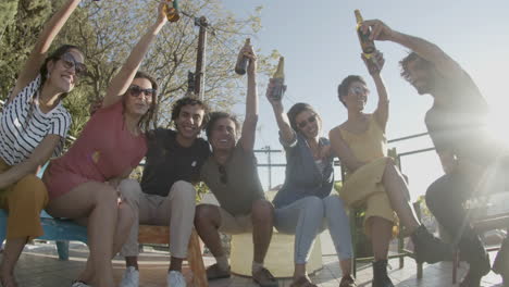 Happy-friends-raising-bottles-of-beer-up-and-looking-at-the-camera-while-sitting-on-rooftop-during-an-outdoor-party