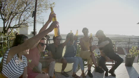 Amigos-Felices-Levantando-Botellas-De-Cerveza-Y-Brindando-Mientras-Se-Sientan-En-La-Azotea-Durante-Una-Fiesta-Al-Aire-Libre