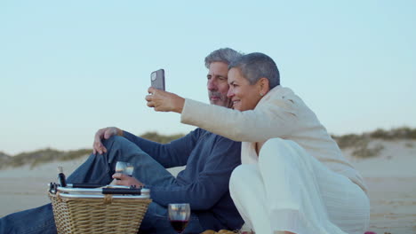 Happy-senior-Caucasian-couple-taking-a-selfie-photo-while-having-a-picnic-on-the-beach