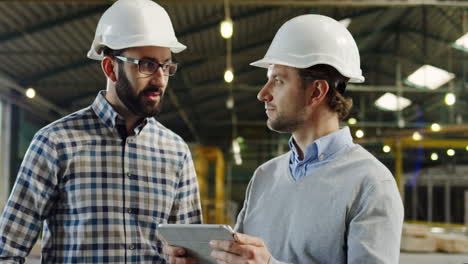 Two-Caucasian-men-wearing-helmets-talking-and-looking-at-the-tablet-screen-in-a-factory,-then-looking-at-the-camera-and-smiling